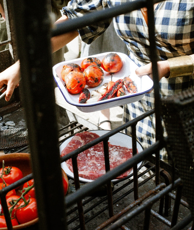 Person holding a tray with roasted tomatoes and other ingredients in an outdoor setting.