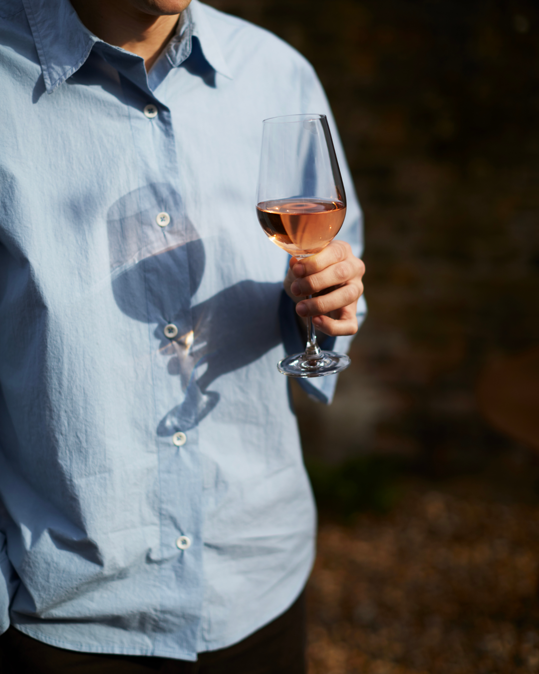 Person in a pale blue shirt holding a glass of rose wine with a blurred background