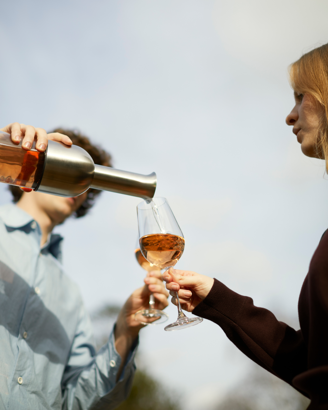 Person pouring rose wine from a stainless satin ETO wine decanter into a glass held by another person against a blue sky