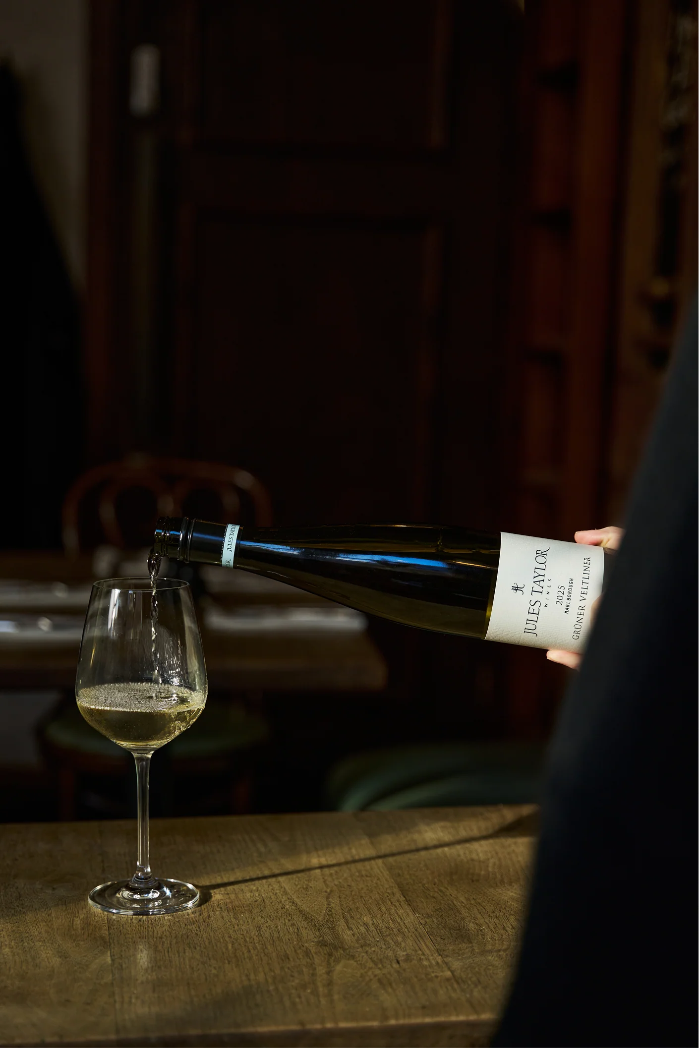 Person pouring white wine from a bottle  of Jules Taylor Gruner Veltliner into a glass on a wooden table.