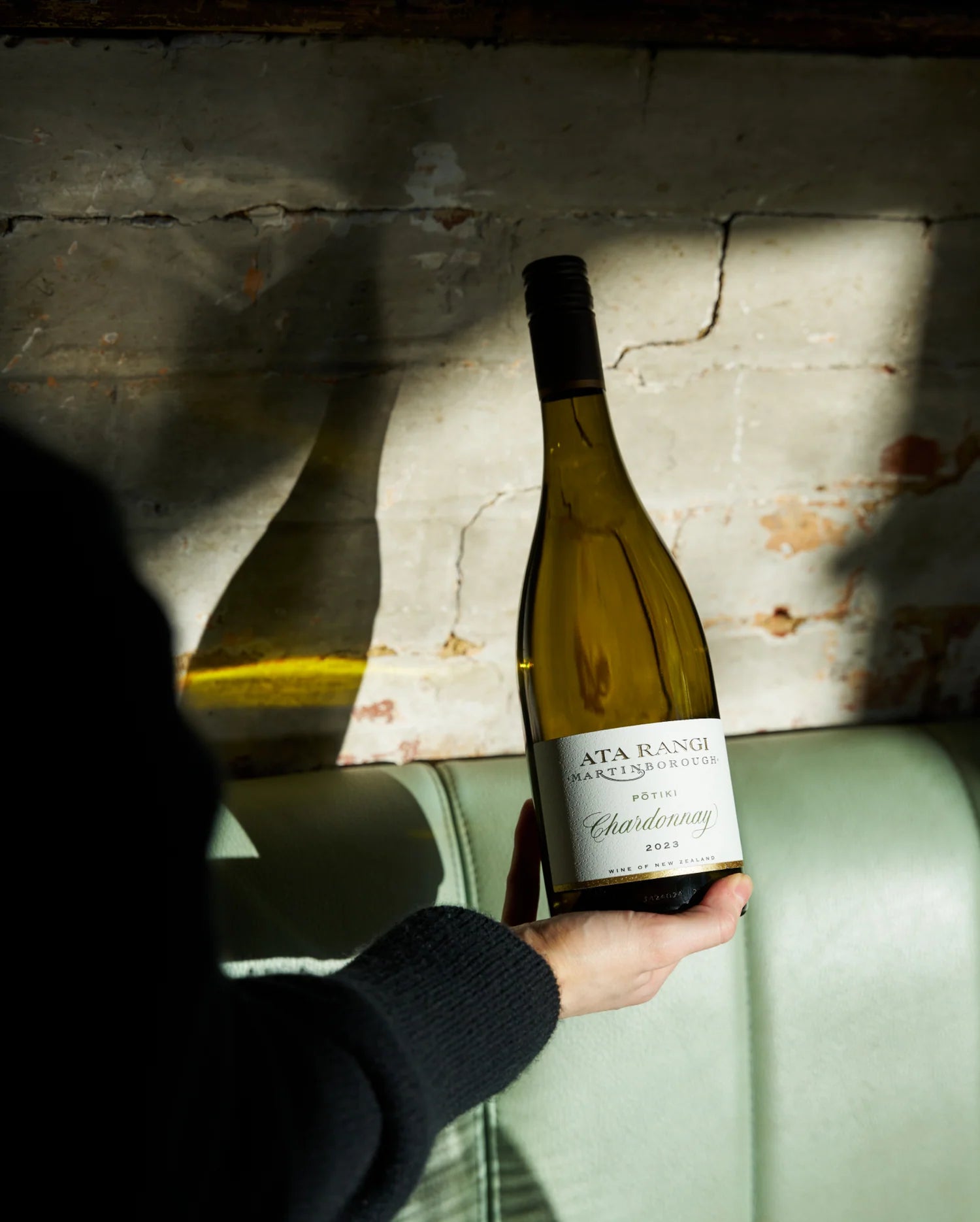 Person holding a bottle of Ata Rangi Chardonnay against a textured wall.