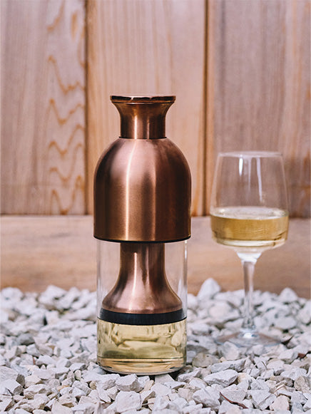 eto wine decanter in copper satin finish filled with white wine sitting on top of a bed of rocks with a glass of white wine at the back