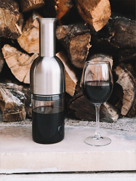 eto wine decanter in stainless satin finish by a stack of logs and a glass of red white